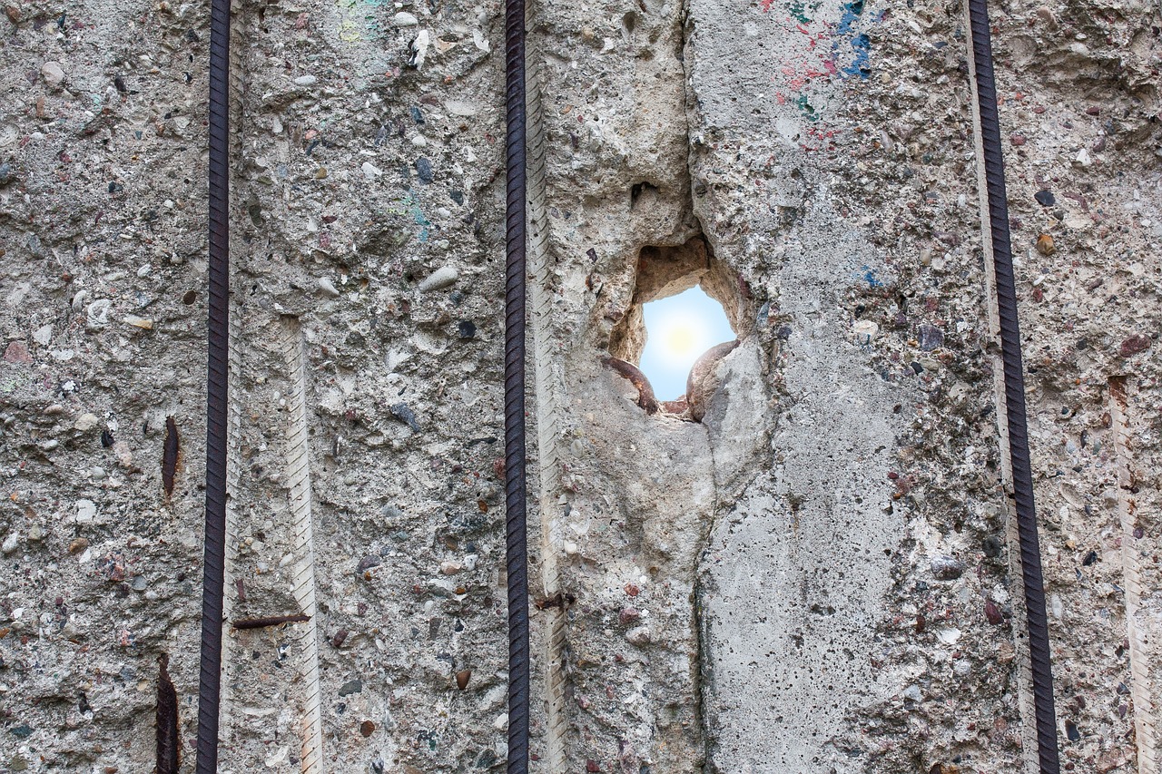 Ein Lichtblick. Eine Betonwand (hier die Berliner Mauer) bröckelt. Ein Loch in der Wand lässt Sonnenstrahlen herein und gibt den Blick auf den blauen Himmel frei.